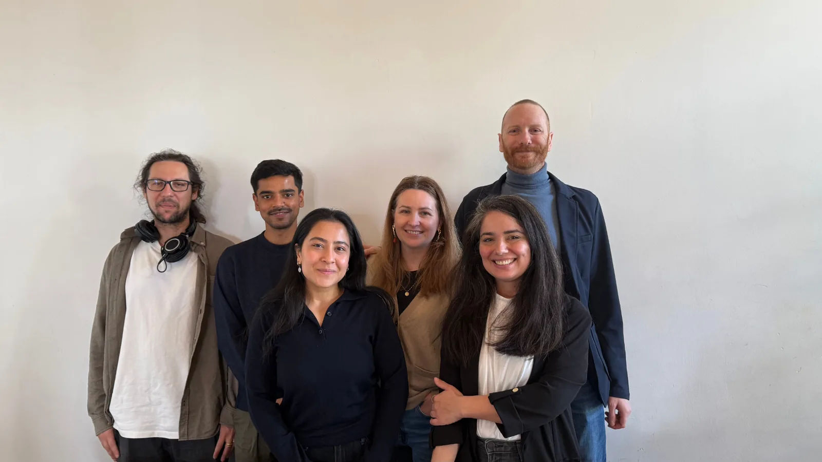 Six people standing and sitting close together in front of a plain pale wall, smiling towards the camera.