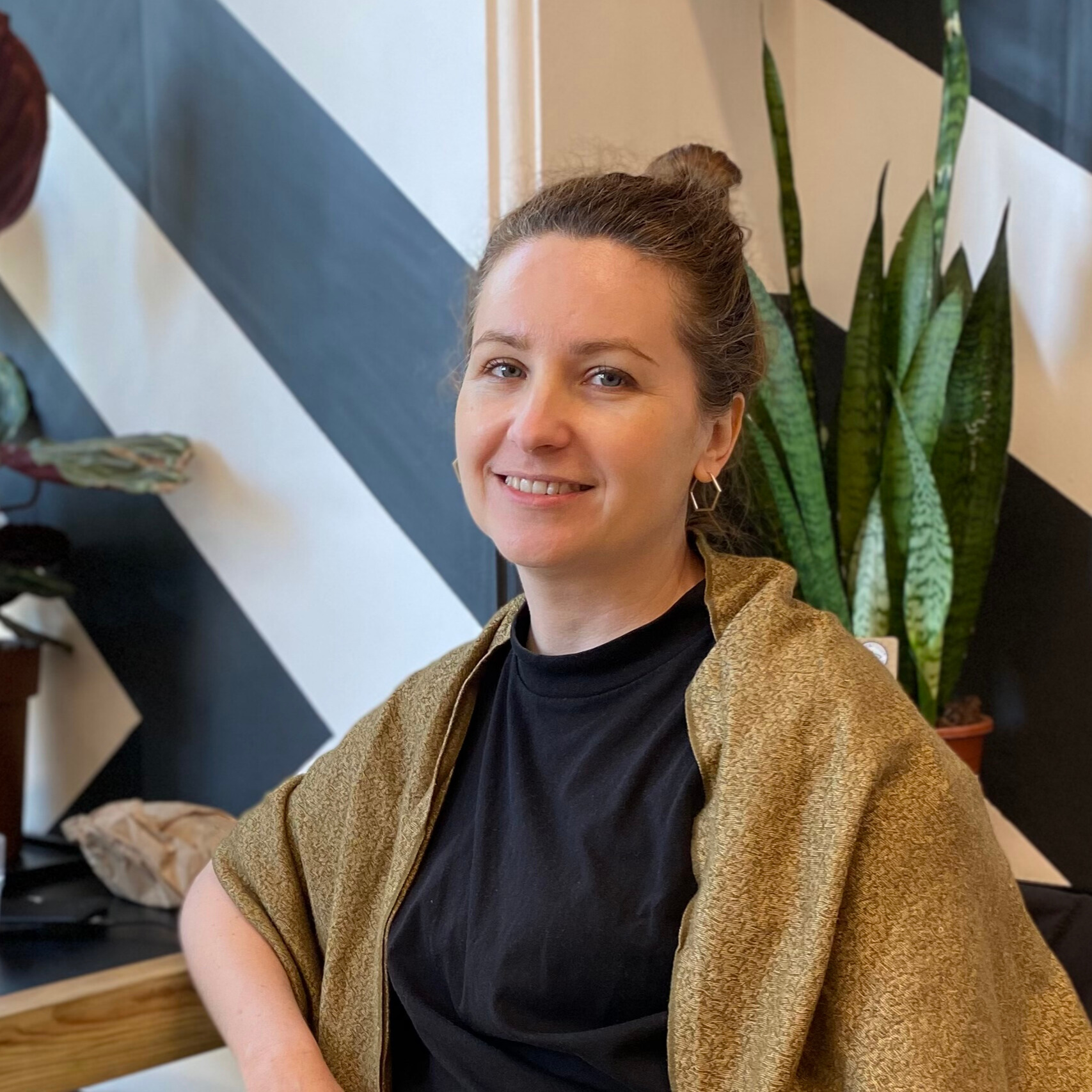 Kata Spychala seated and smiling, an ochre shawl over a black shirt, a plant behind her